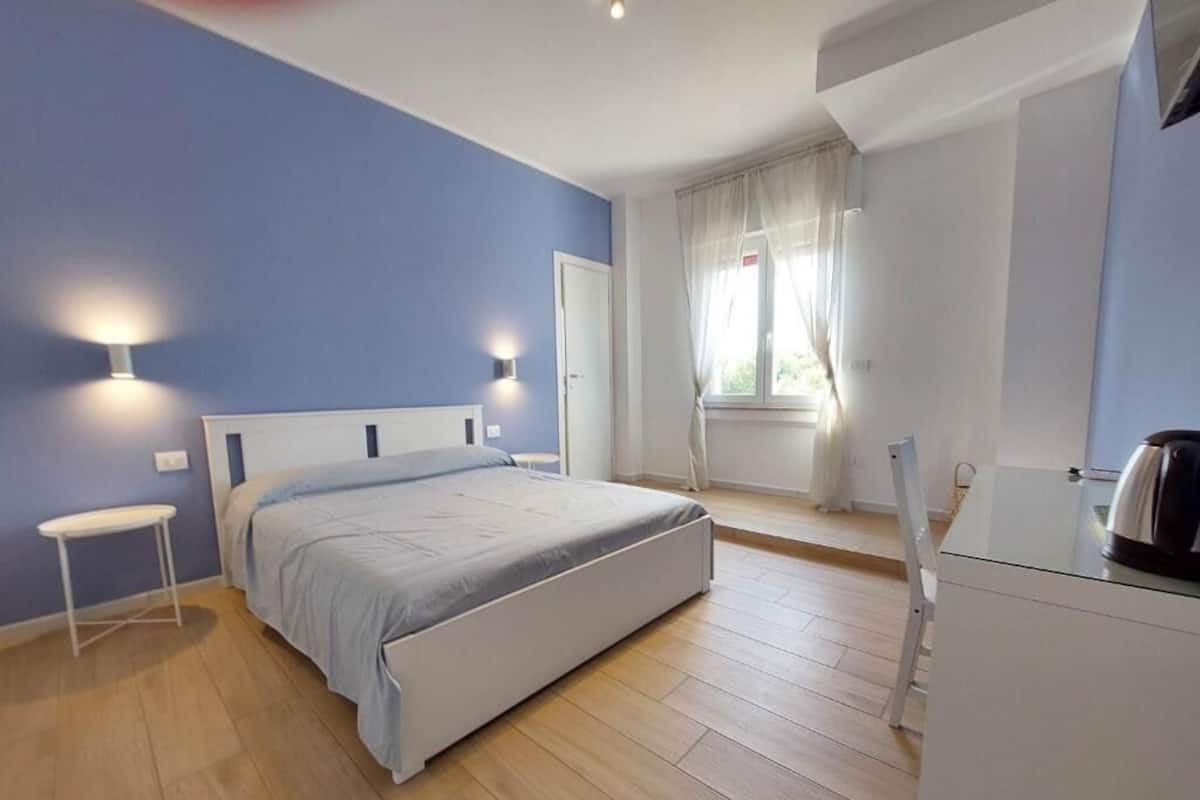 Un dormitorio con cama, mesitas de noche, ventana con cortinas y una pequeña cocina.