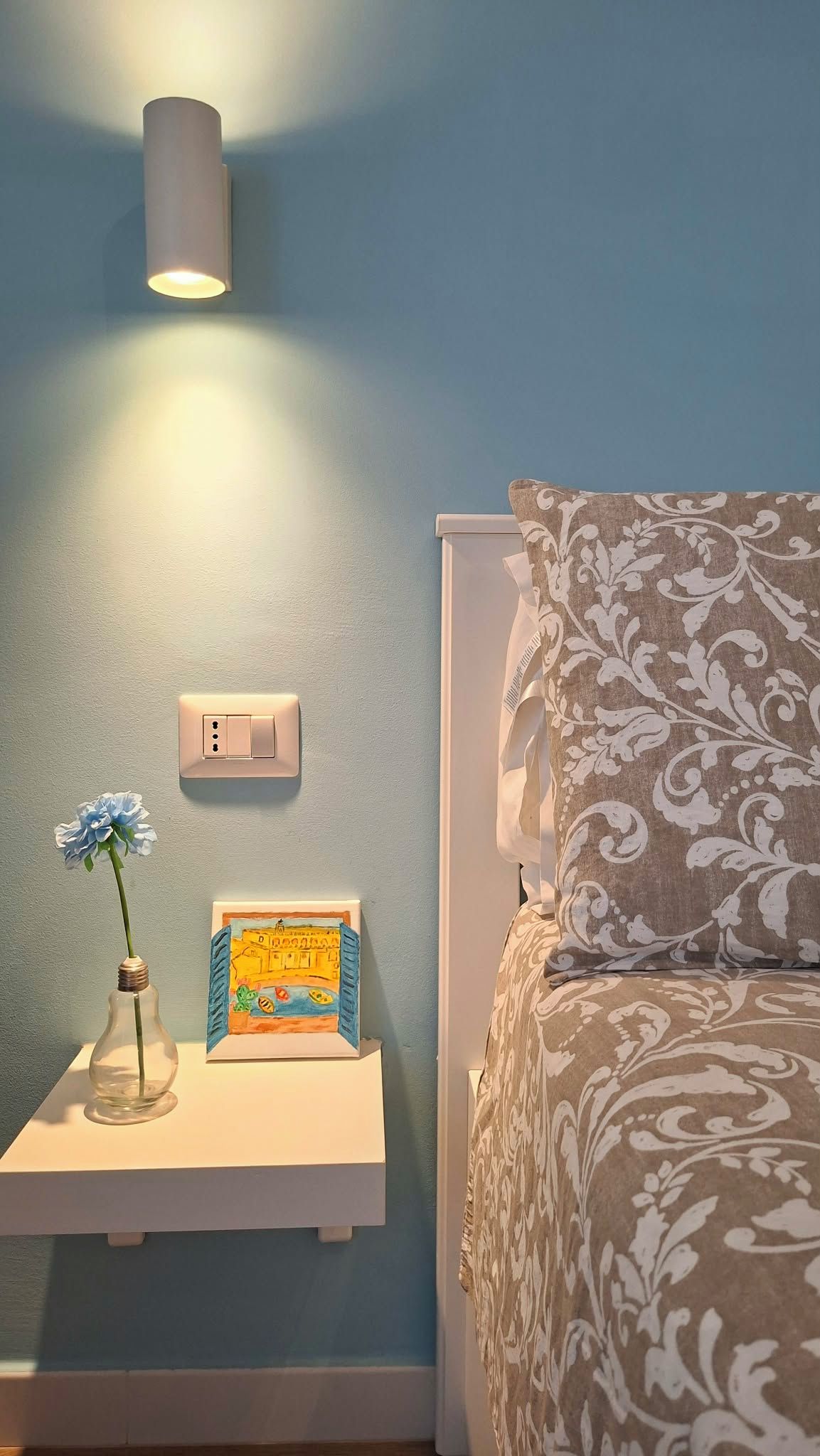 Ein Schlafzimmer mit einem Bett, einem Nachttisch mit Lampe, einer Steckdose und einem gerahmten Bild.