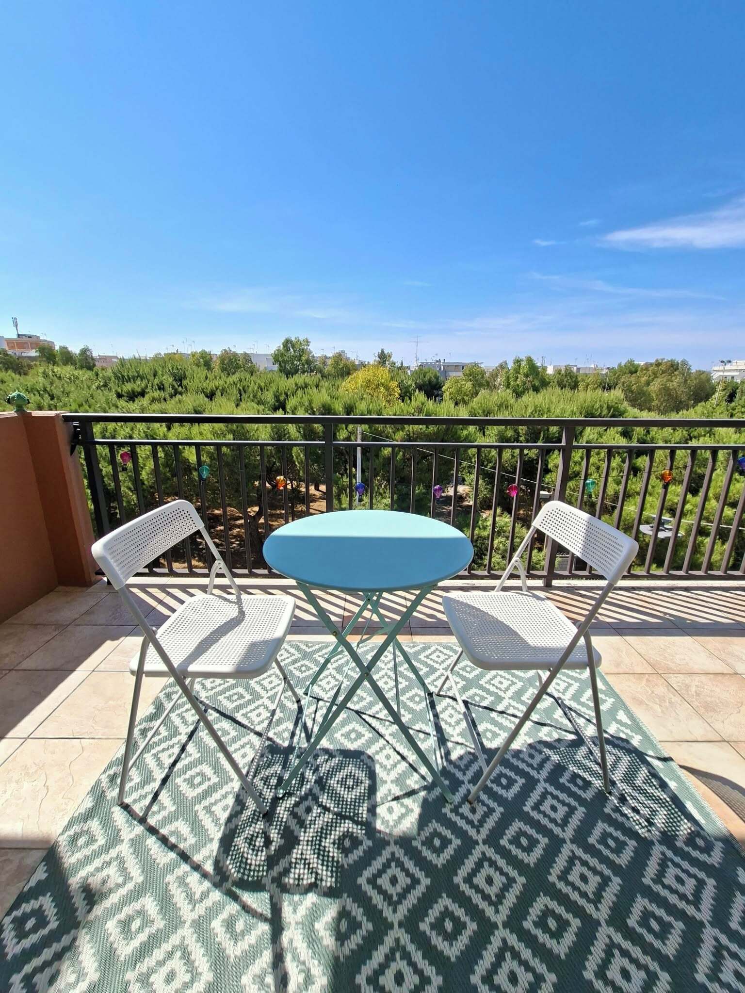 A balcony with a table and two chairs, a patterned rug, and a view of greenery and buildings.