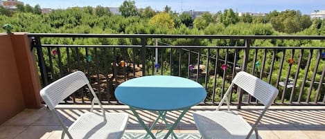 A balcony with a table and two chairs, a patterned rug, and a view of greenery and buildings.