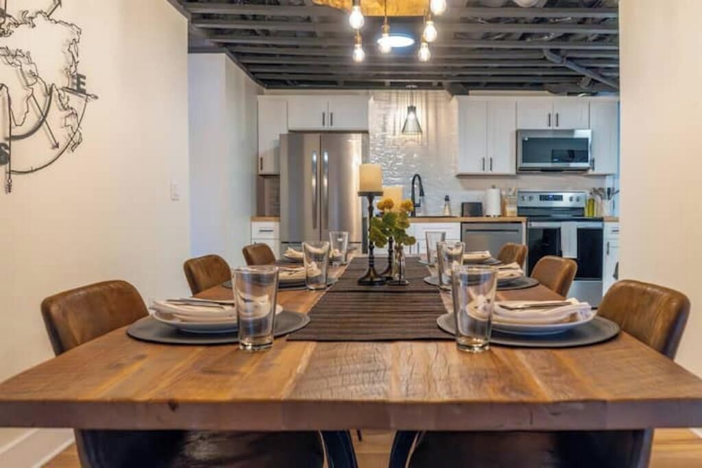 Uma sala de jantar com uma mesa de madeira posta para seis pessoas, cercada por cadeiras. Uma cozinha com eletrodomésticos modernos é visível ao fundo.