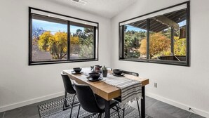 Un'area pranzo con tavolo apparecchiato per quattro persone, ampie finestre con cornice nera e vista su alberi e un edificio.