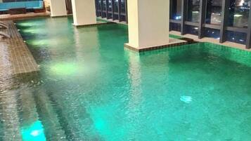 An indoor swimming pool with clear water, tiled flooring, and a view of the city at night through large windows.