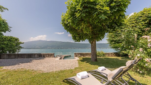 Ein Poolbereich mit Liegestühlen, einem Baum und Blick auf einen See und Berge.