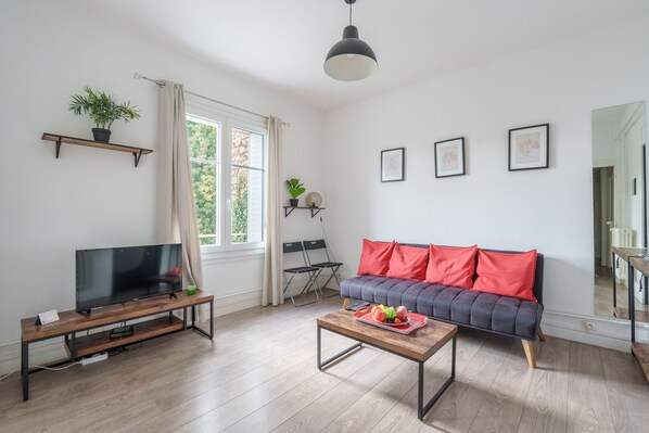 A living room with a TV on a wooden stand, a sofa with red cushions, a coffee table with fruit, and a large window with curtains.