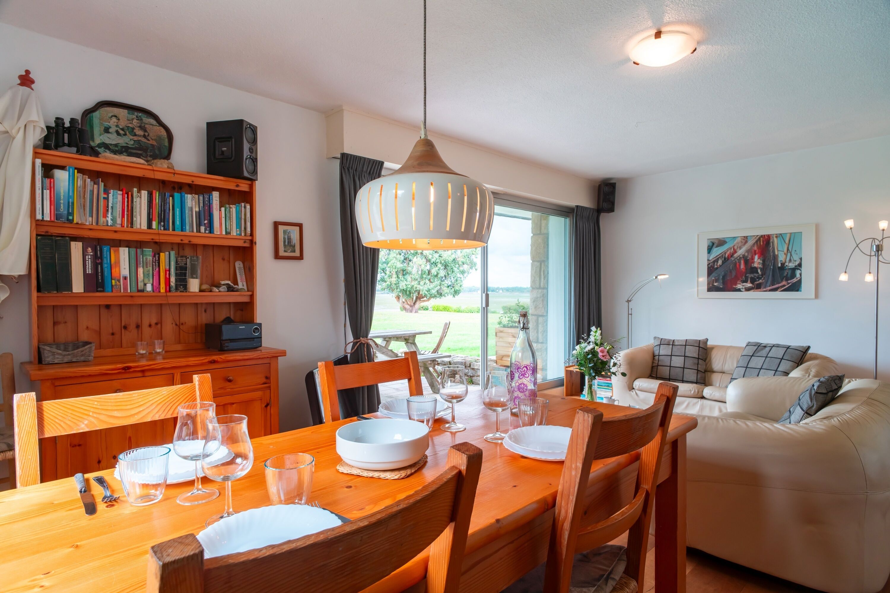 Ein Essbereich mit einem Holztisch, gedeckt für vier Personen, umgeben von Stühlen. Eine Bücherwand mit Büchern ist links, und ein Sofa im Hintergrund.