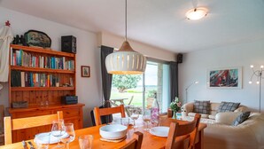 Un coin repas avec une table en bois dressée pour quatre personnes, entourée de chaises. Une bibliothèque remplie de livres se trouve à gauche, et un canapé est visible au fond.