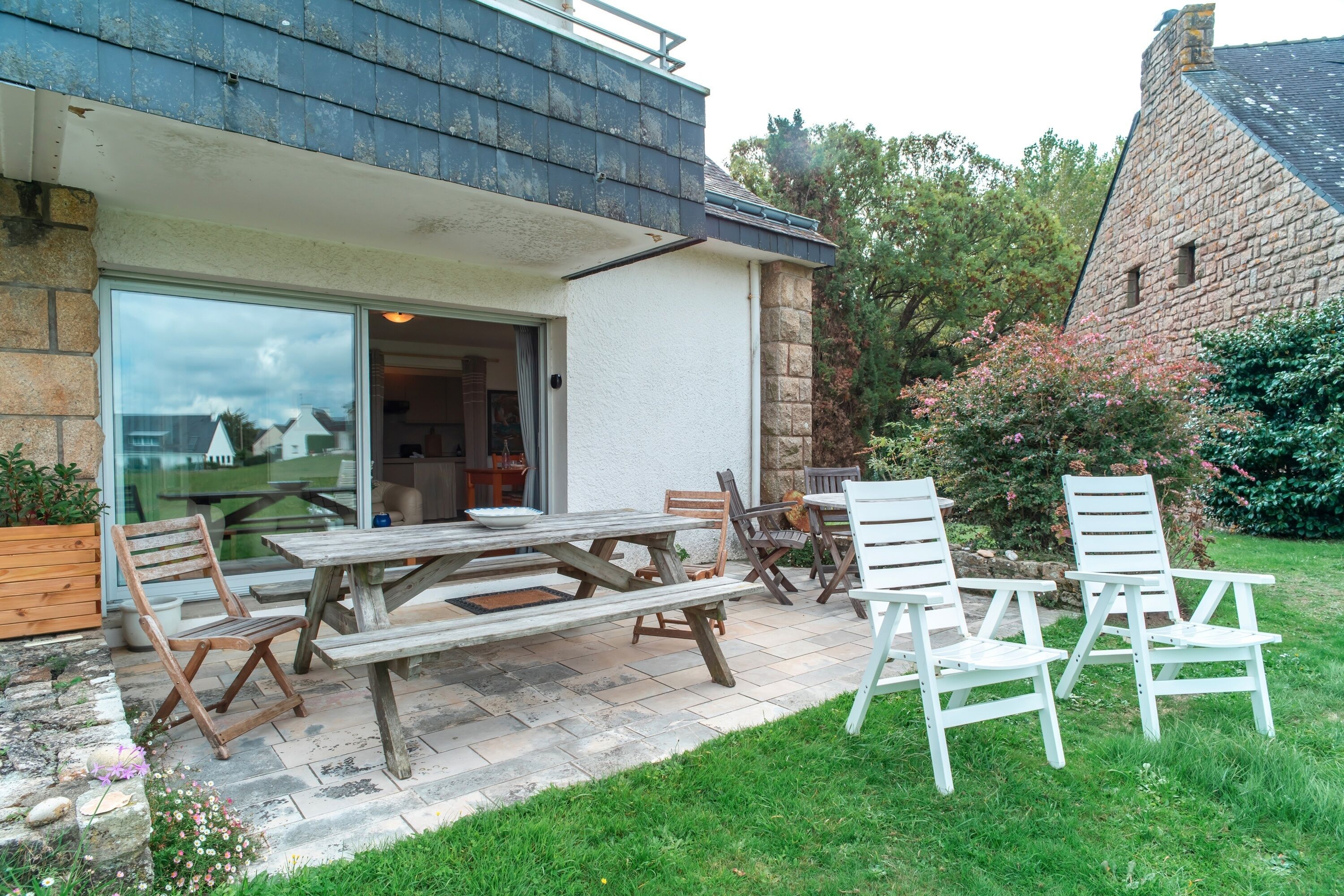 An outdoor patio with wooden benches and chairs, a stone wall, and a garden area with flowers and shrubs.