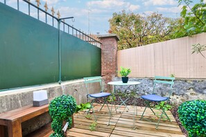 Une terrasse en bois avec deux chaises vertes et une petite table, entourée de verdure et d’un mur de briques.