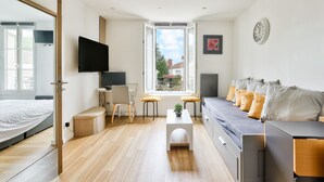 A modern, well-lit room with a bed, a sofa, a desk with a chair, a television, and a window with a view of the outside.