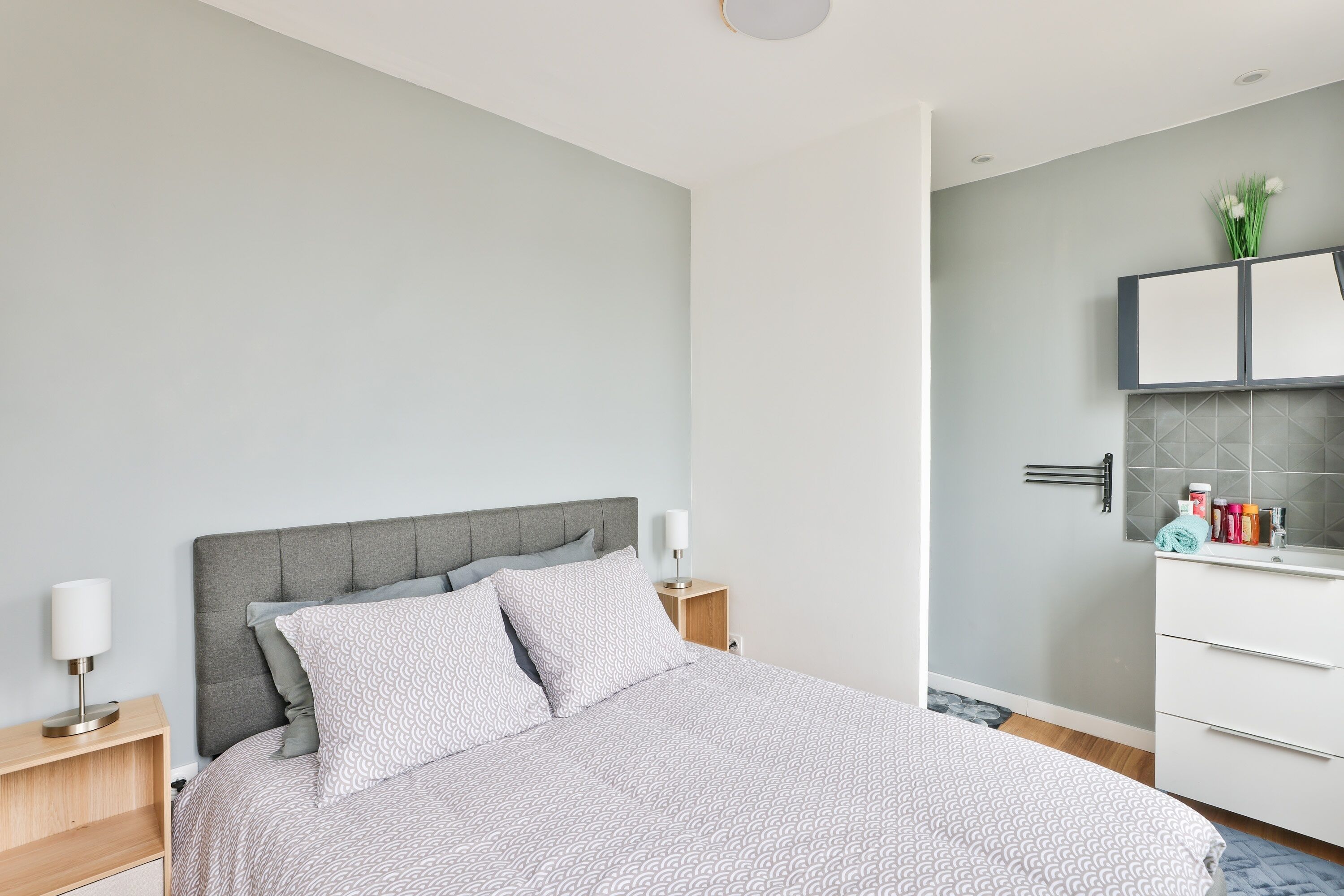 A modern bedroom with a grey upholstered headboard, a neatly made bed with patterned bedding, a bedside table with a lamp, and a bathroom with a white vanity and tiled backsplash.