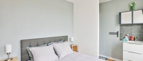 A modern bedroom with a grey upholstered headboard, a neatly made bed with patterned bedding, a bedside table with a lamp, and a bathroom with a white vanity and tiled backsplash.