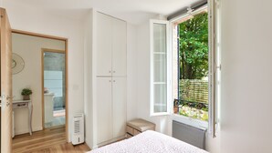 A bedroom with a bed, a wardrobe, a radiator, a window with a view of greenery, and a door leading to a bathroom.