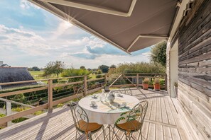 A wooden deck with a round table and chairs, surrounded by potted plants and a view of the countryside.
