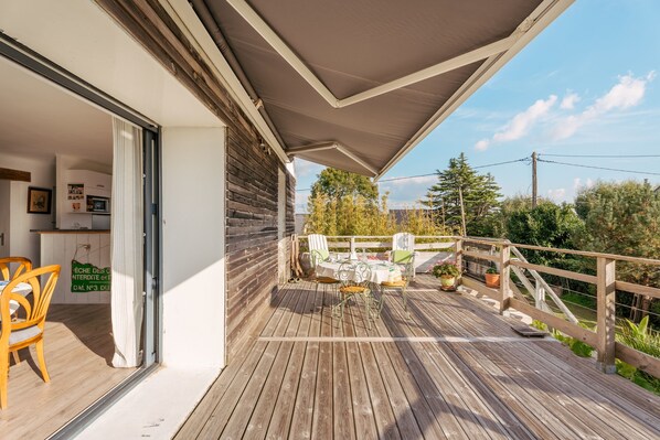 A wooden deck with a table and chairs, a covered area, and a view of trees and a building in the background.