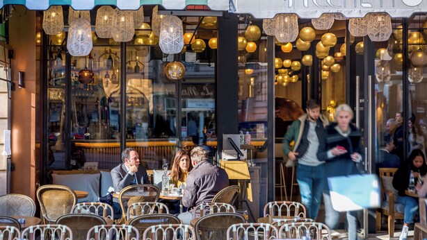 Ein Straßencafé mit Korbstühlen und Tischen, einem Streifenmarkisen und hängenden Lichterketten.