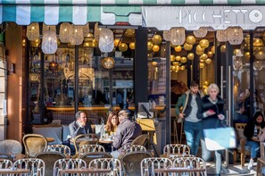 Un café al aire libre con sillas y mesas de mimbre, un toldo a rayas y luces decorativas colgantes.