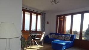 Ein Wohnzimmer mit einem blauen Sofa, einem Esstisch mit Stühlen und großen Fenstern.
