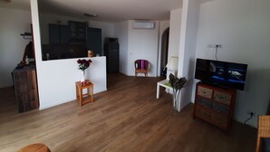 Ein Wohnzimmer mit Holzboden, einem Fernseher, einem kleinen Tisch mit einer Vase und einer Küchenzeile mit Schränken und Geräten.