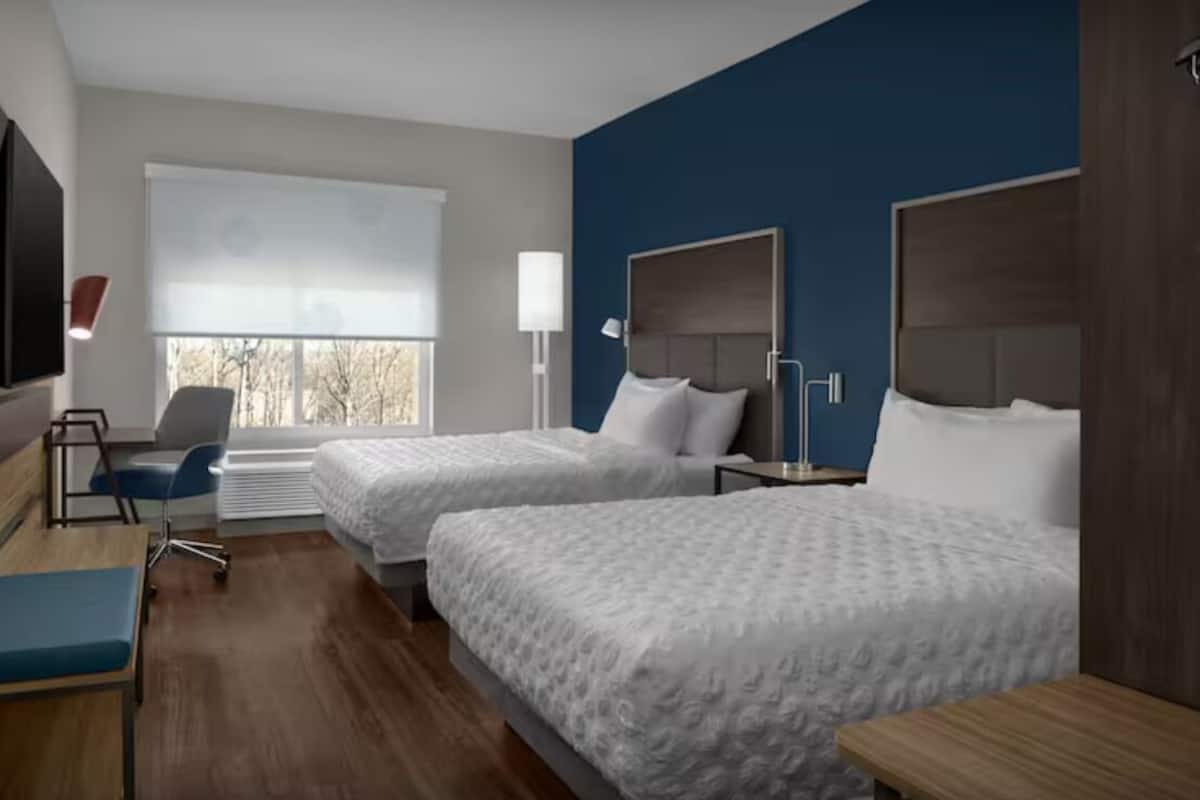 A hotel room with two beds, a desk, a chair, a TV, and a window with blinds.