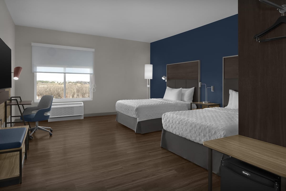 A hotel room with two beds, a desk, a chair, a lamp, and a large window.