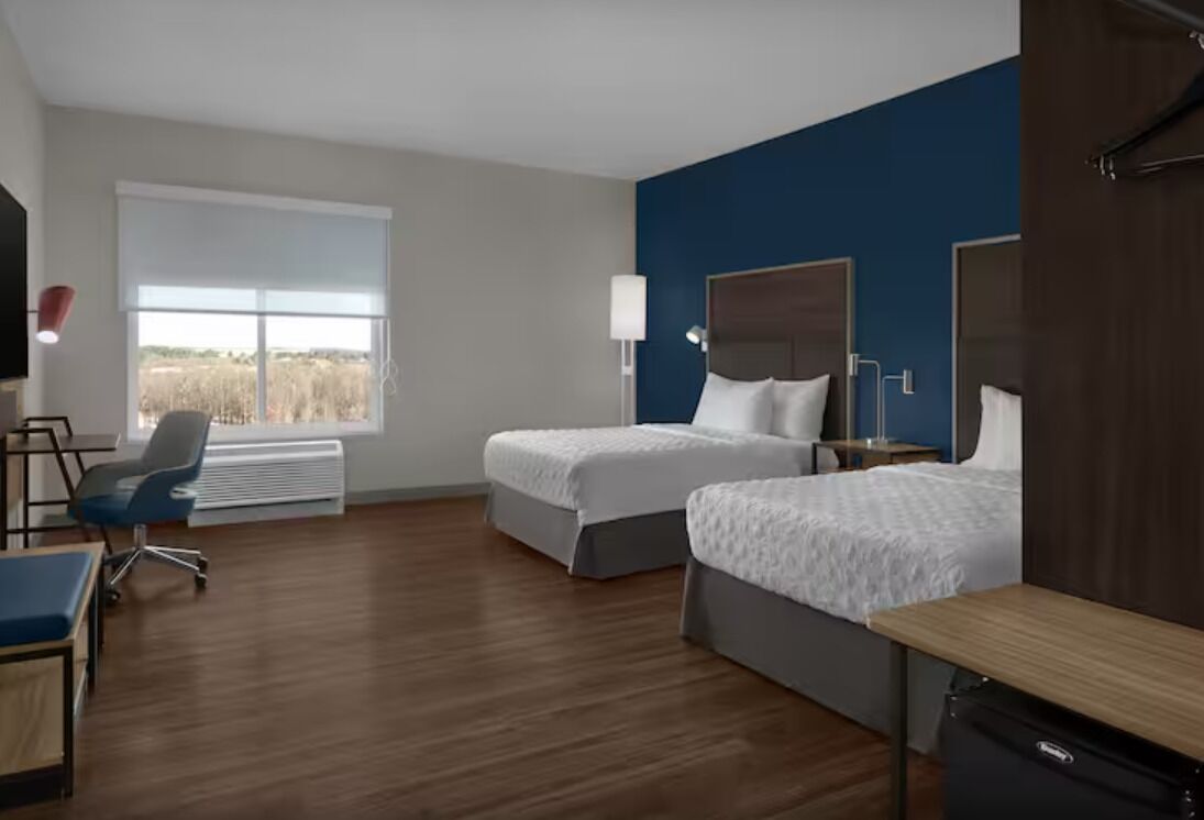 A hotel room with two beds, a desk, a chair, a lamp, and a large window.