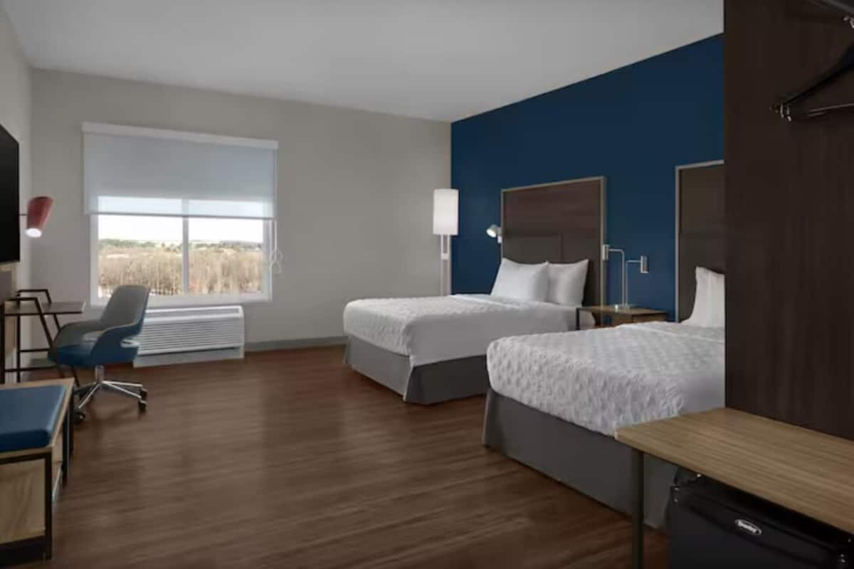 A hotel room with two beds, a desk, a chair, a lamp, and a large window.