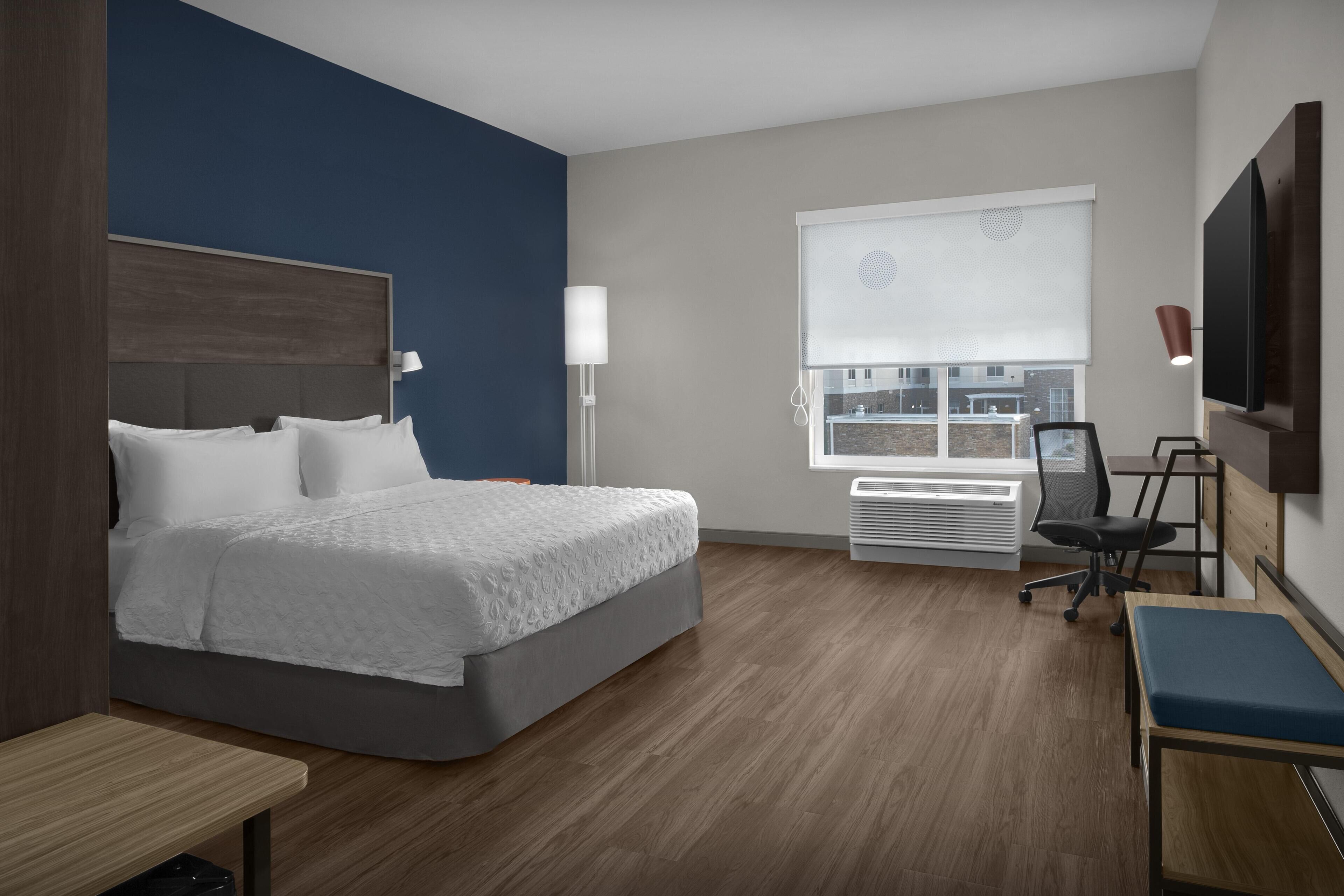 A modern hotel room with a large bed, a desk, a chair, and a window with a view of buildings.