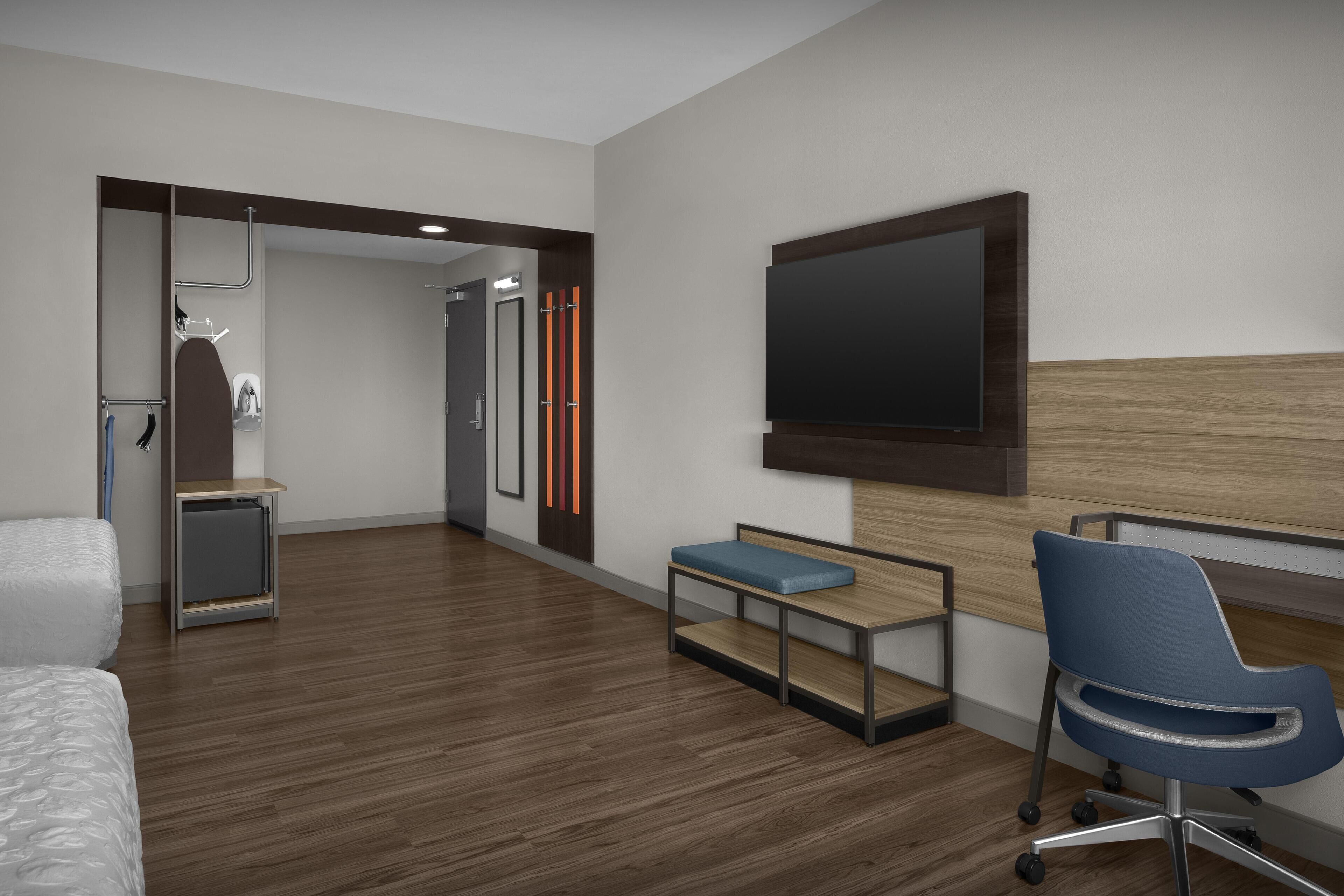 A modern hotel room with a wooden floor, a bed, a desk with a chair, a TV mounted on the wall, and a shower area.
