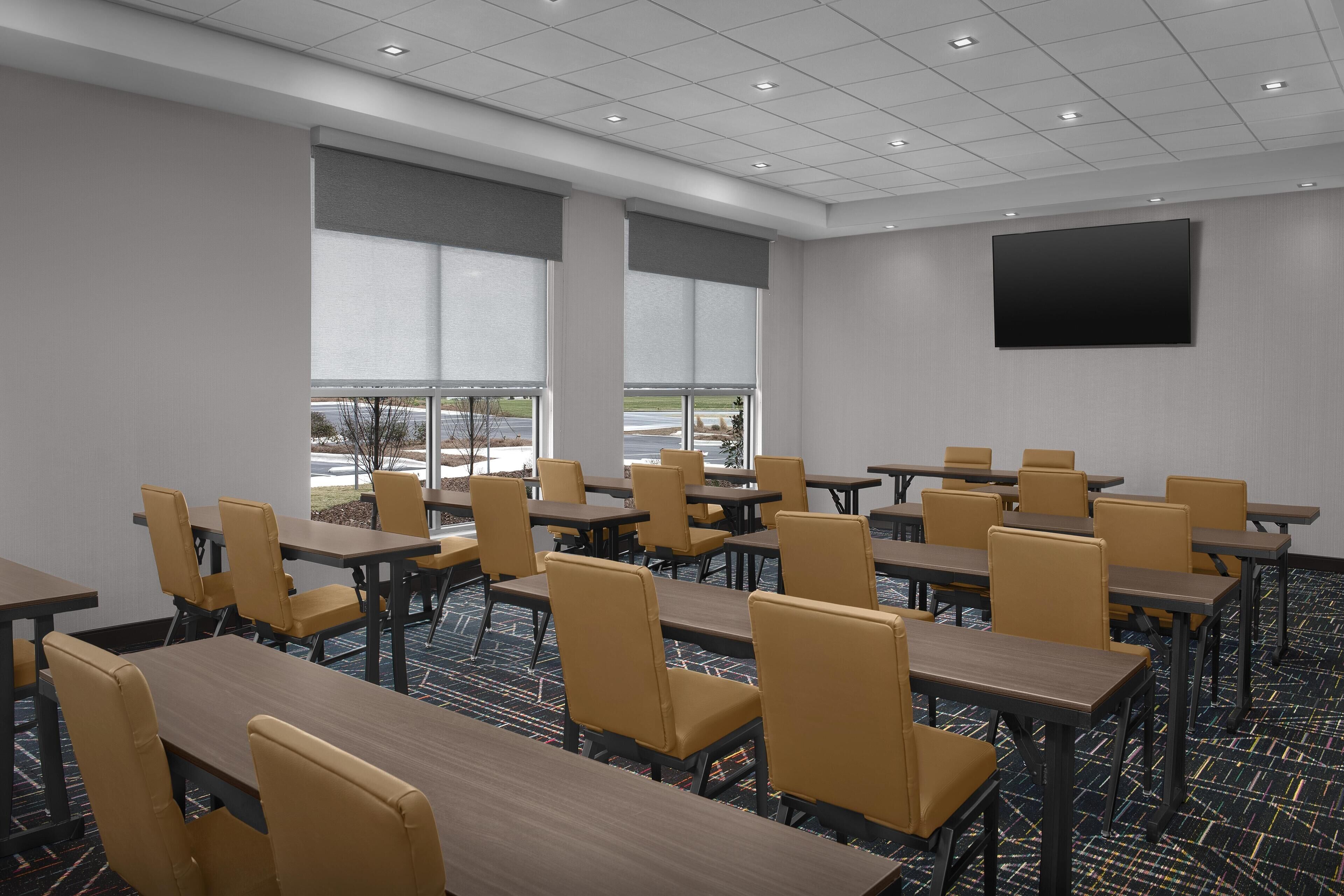 A conference room with tables and chairs, large windows, and a flat-screen TV on the wall.