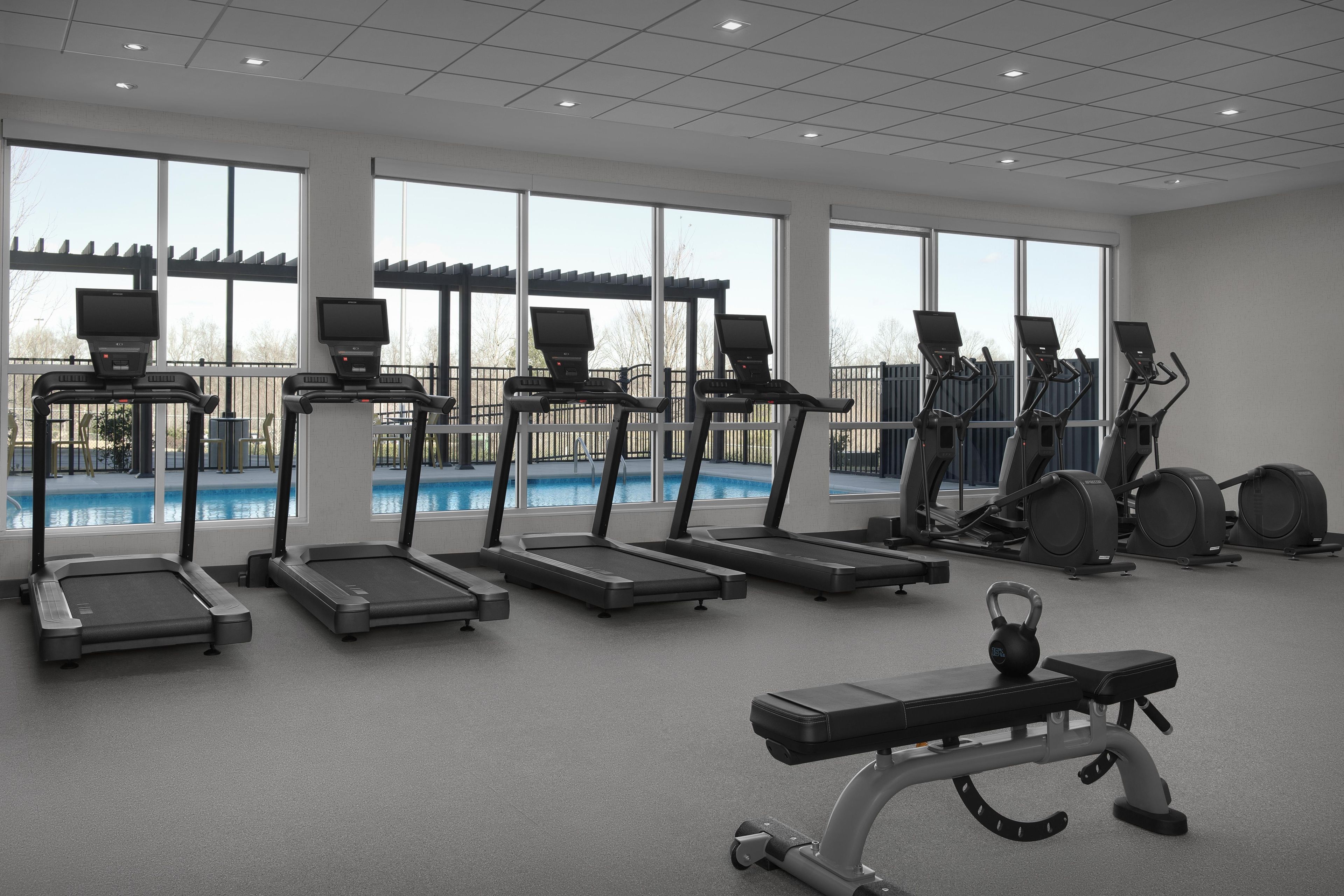 A gym with multiple treadmills, a bench, and a rowing machine, with large windows overlooking a swimming pool.