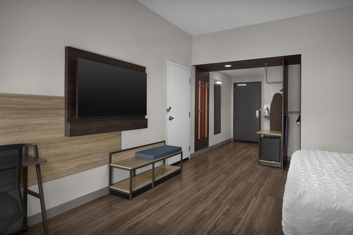 A modern hotel room with a bed, a desk, a bench, a TV, and a door leading to a bathroom.