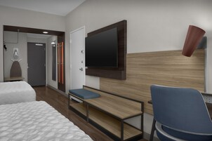 Habitación de hotel con cama, escritorio con silla, televisor en la pared y zona de ducha.