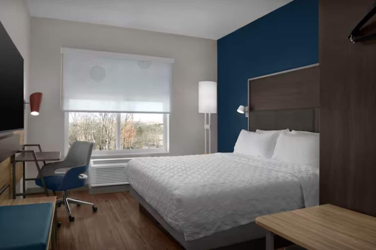A hotel room with a bed, a desk, a chair, a window with blinds, and a lamp.
