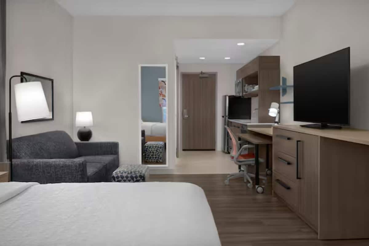 A modern hotel room with a bed, a desk, a TV, and a seating area.