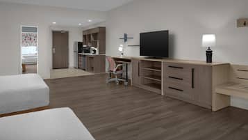 A modern hotel room with a wooden floor, a desk with a flat-screen TV, a chair, a bed, and a kitchenette.