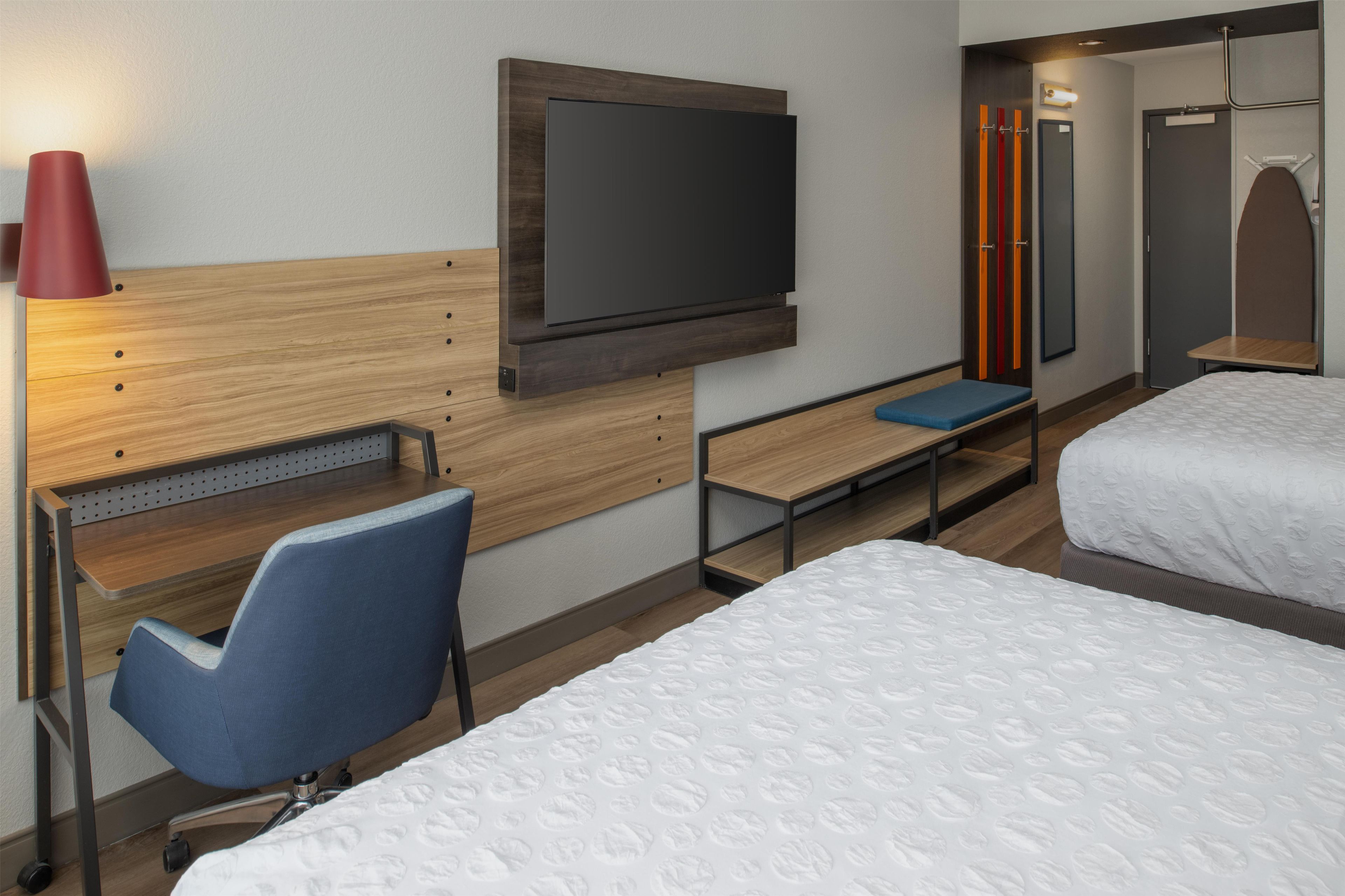 A hotel room with a bed, a desk with a chair, a television mounted on the wall, and a wardrobe.