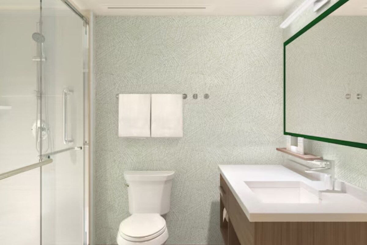 A bathroom with a glass shower enclosure, a white toilet, a white sink with a wooden vanity, and a mirror with a green frame.