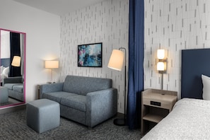 A hotel room with a grey sofa, a small ottoman, a bed, a nightstand with a lamp, a wall-mounted painting, and a mirror with a pink frame. - Home2 Suites By Hilton Salisbury Fruitland (Fruitland)