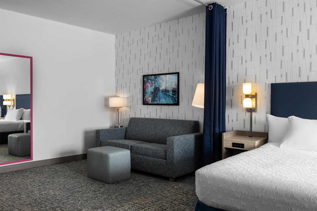 A hotel room with a gray sofa, a small ottoman, a bed with white bedding, a nightstand with a lamp, a wall-mounted TV, and a mirror with a pink frame.