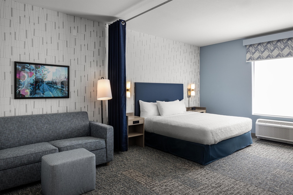 A hotel room with a bed, a sofa, a small table, a lamp, and a painting on the wall.