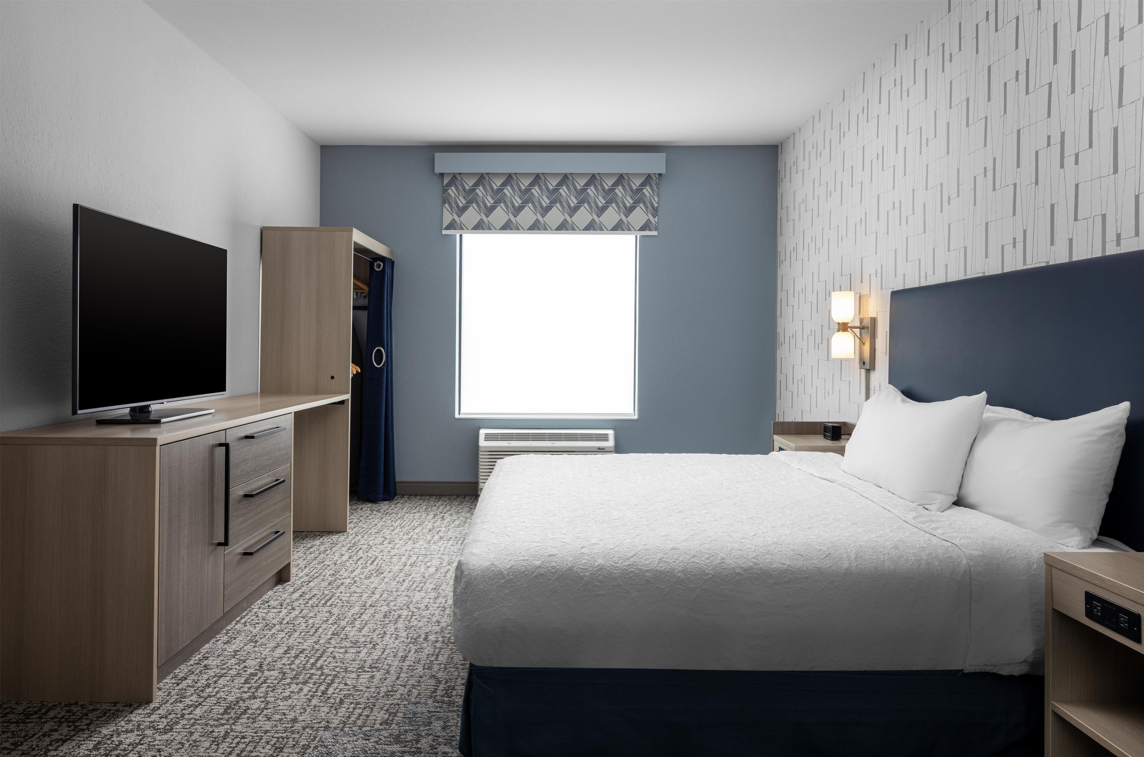 A hotel room with a bed, a TV, a wardrobe, a window with a patterned valance, and a wall-mounted lamp.