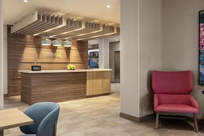 A hotel reception area with a wooden reception desk, a blue chair, and a red chair. - Home2 Suites By Hilton Salisbury Fruitland (Fruitland)