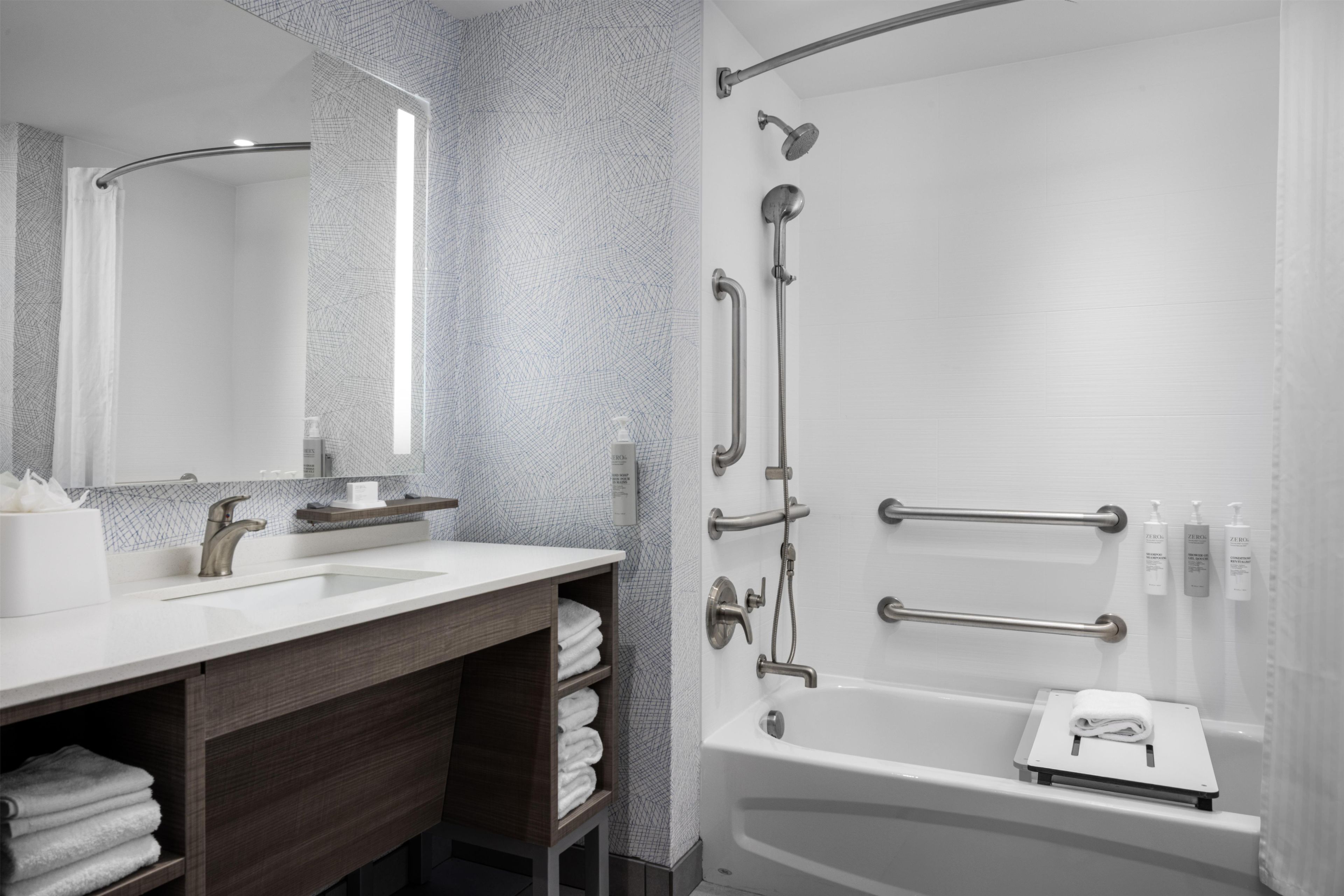 A modern bathroom with a large mirror, a white sink, a bathtub with grab bars, and a towel rack.