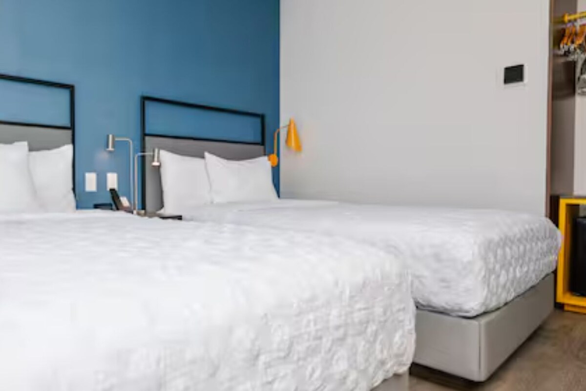 A hotel room with two beds, each with white bedding, a blue wall, and a small table with a lamp.