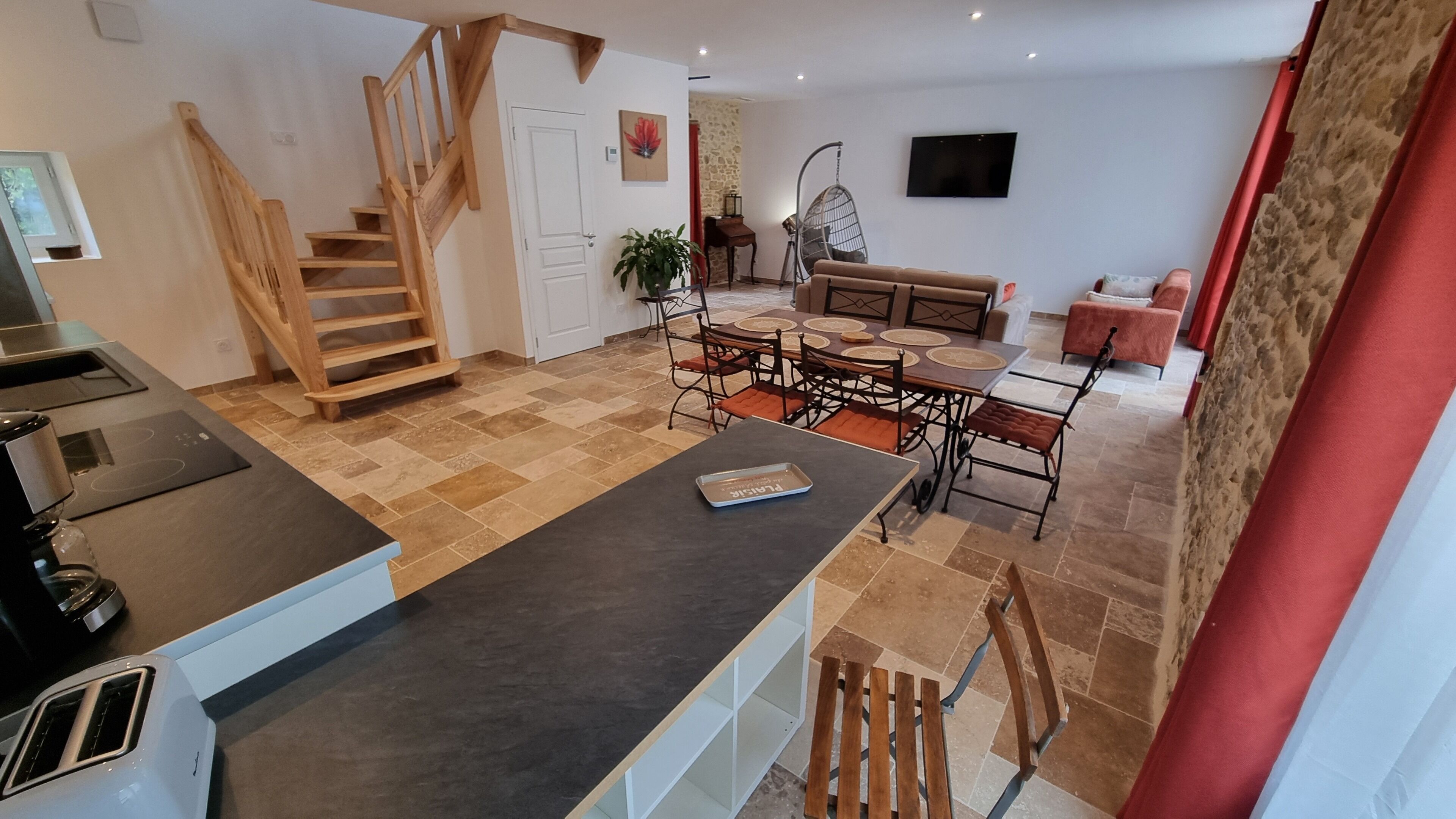 A modern kitchen with a wooden staircase, a dining area with a round table and chairs, and a living area with a sofa and a TV.