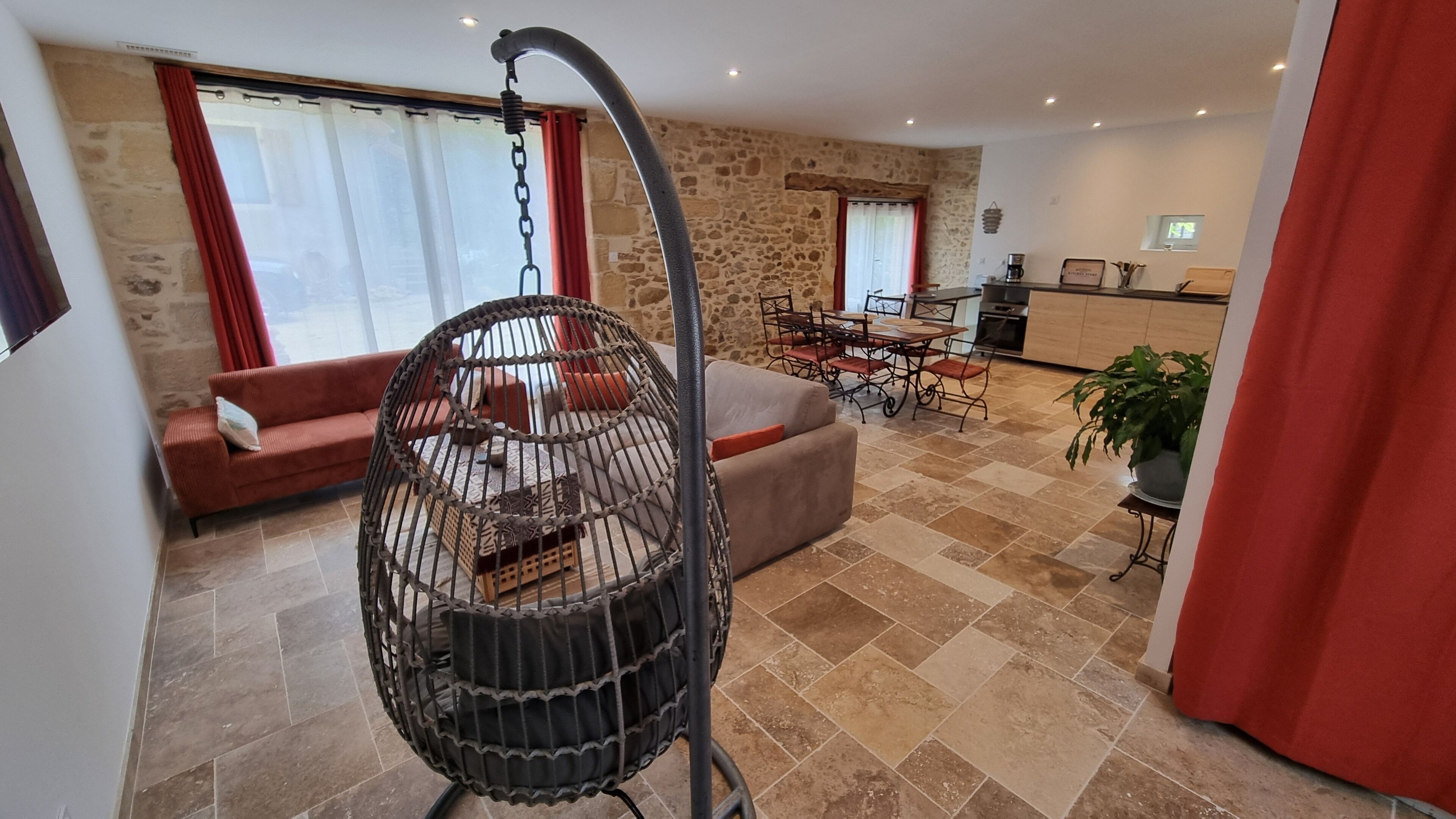 A living room with a hanging chair, a sofa, a dining table with chairs, a kitchen area, and a stone wall.