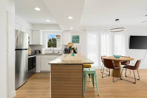 A modern kitchen with white cabinets, a stainless steel refrigerator, a wooden island, and a dining area with a table and chairs.