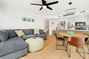 A modern living room with a gray sectional sofa, a round ottoman, a dining table with chairs, and a kitchen area in the background.