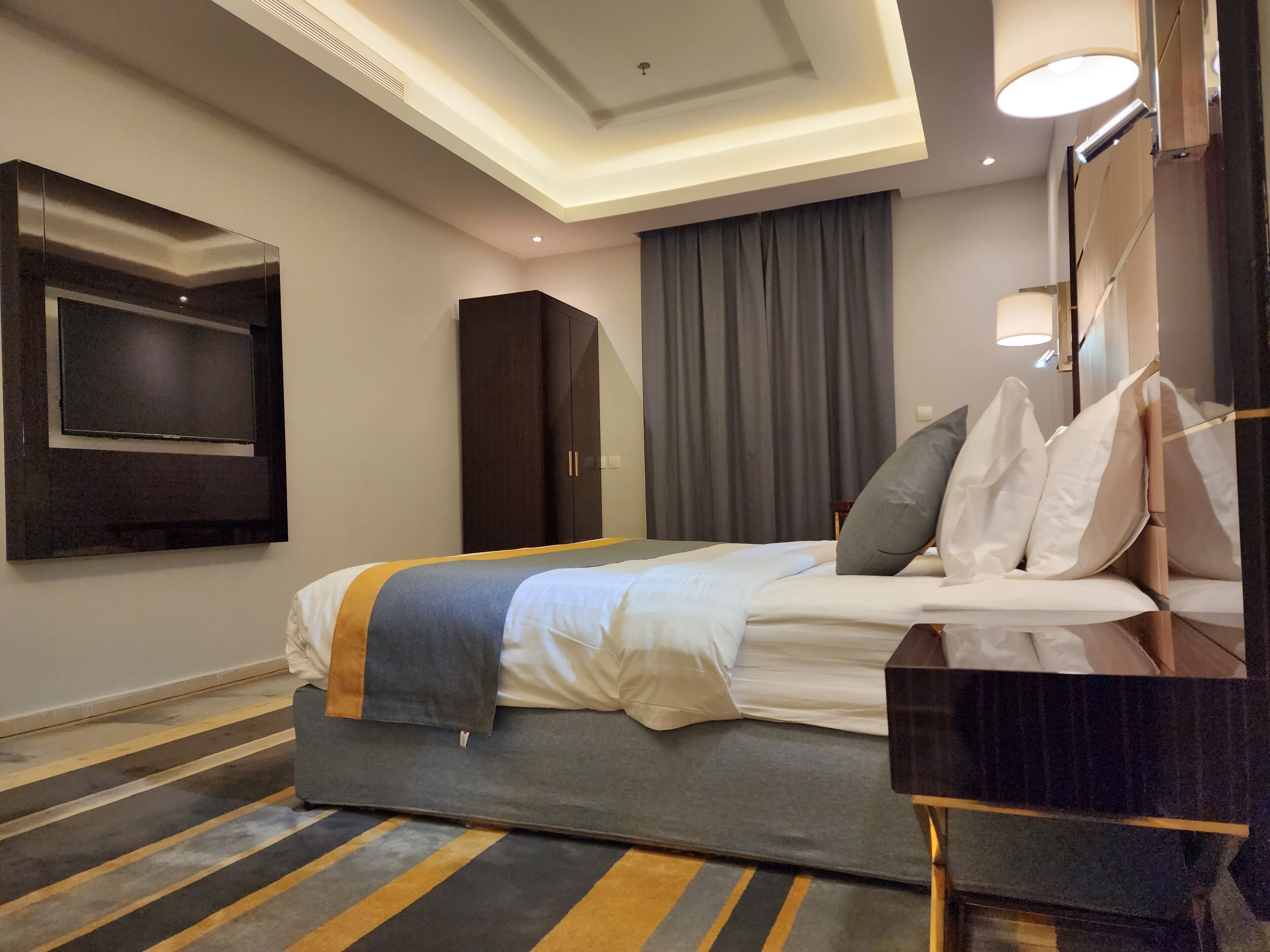 Ein Hotelzimmer mit Bett, einem an der Wand montierten Fernseher, einem Kleiderschrank und einem Schreibtisch mit Lampe.